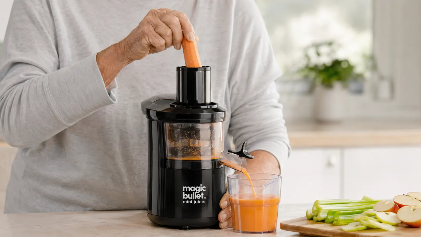 An older woman's hands feeding a carrot into the narrow feed chute of a small countertop juicer