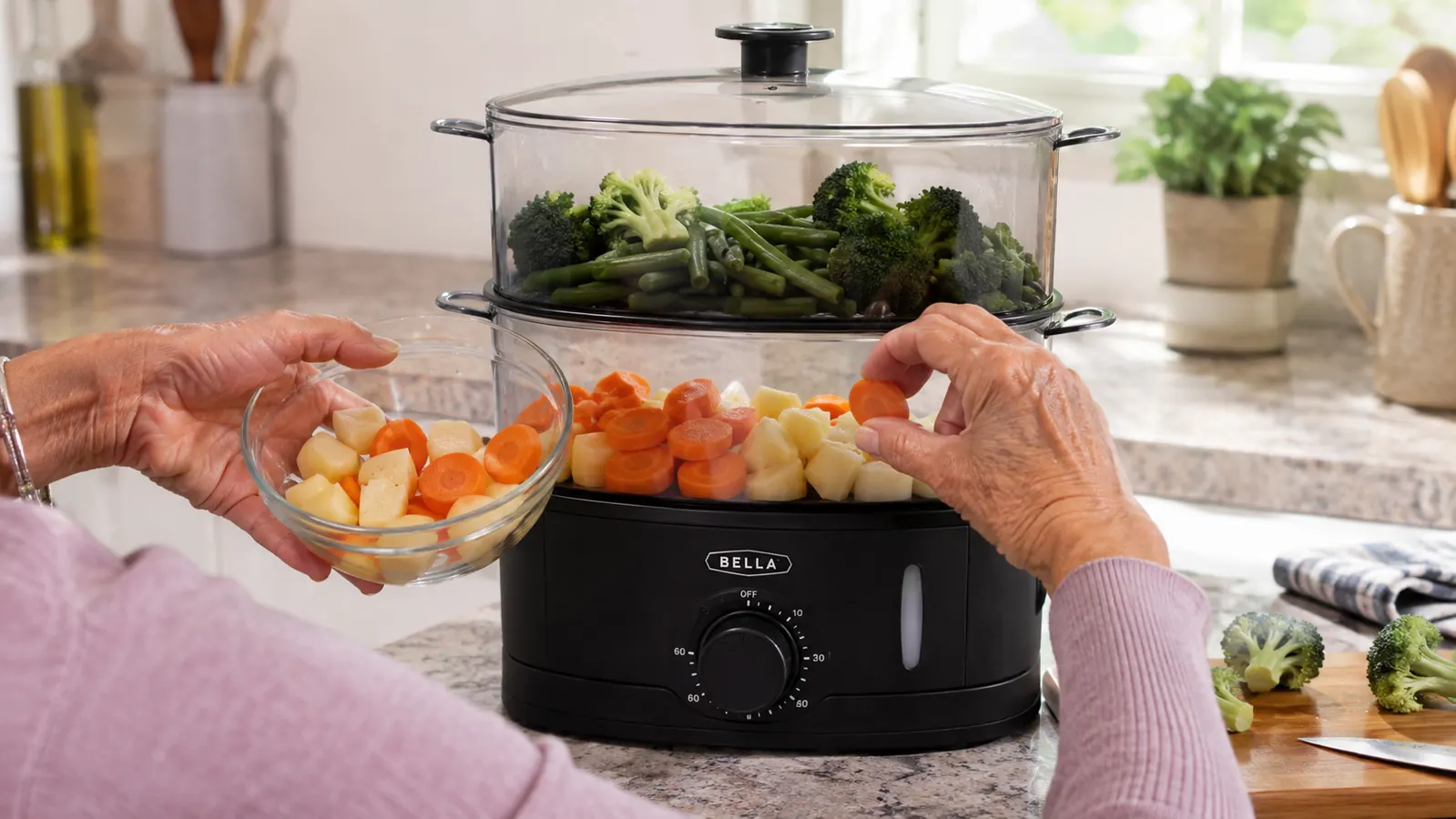 Hands loading cut vegetables into the two stacked baskets of an electric steamer, root vegetables in the lower basket and delicate vegetables in the upper basket